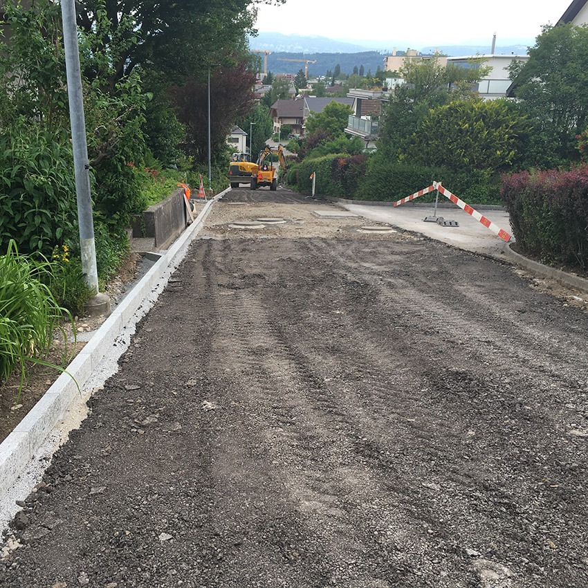 Straßenbauarbeiten in Wohngebiet, Baustellenfahrzeug auf unbefestigtem Weg, Absperrung im Hintergrund, grüne Umgebung.