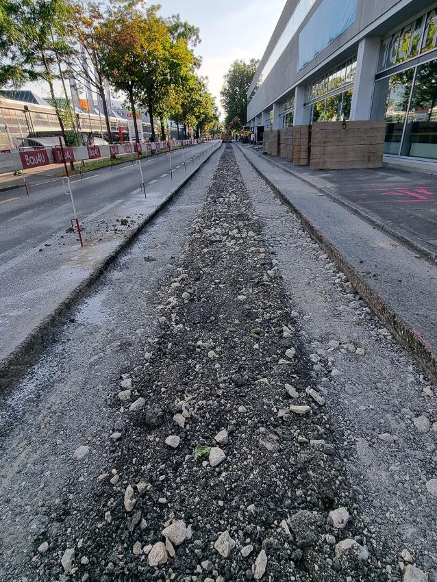 Straßenbaustelle mit offenem Graben, links Absperrungen, daneben ein Gebäude bei Tageslicht.